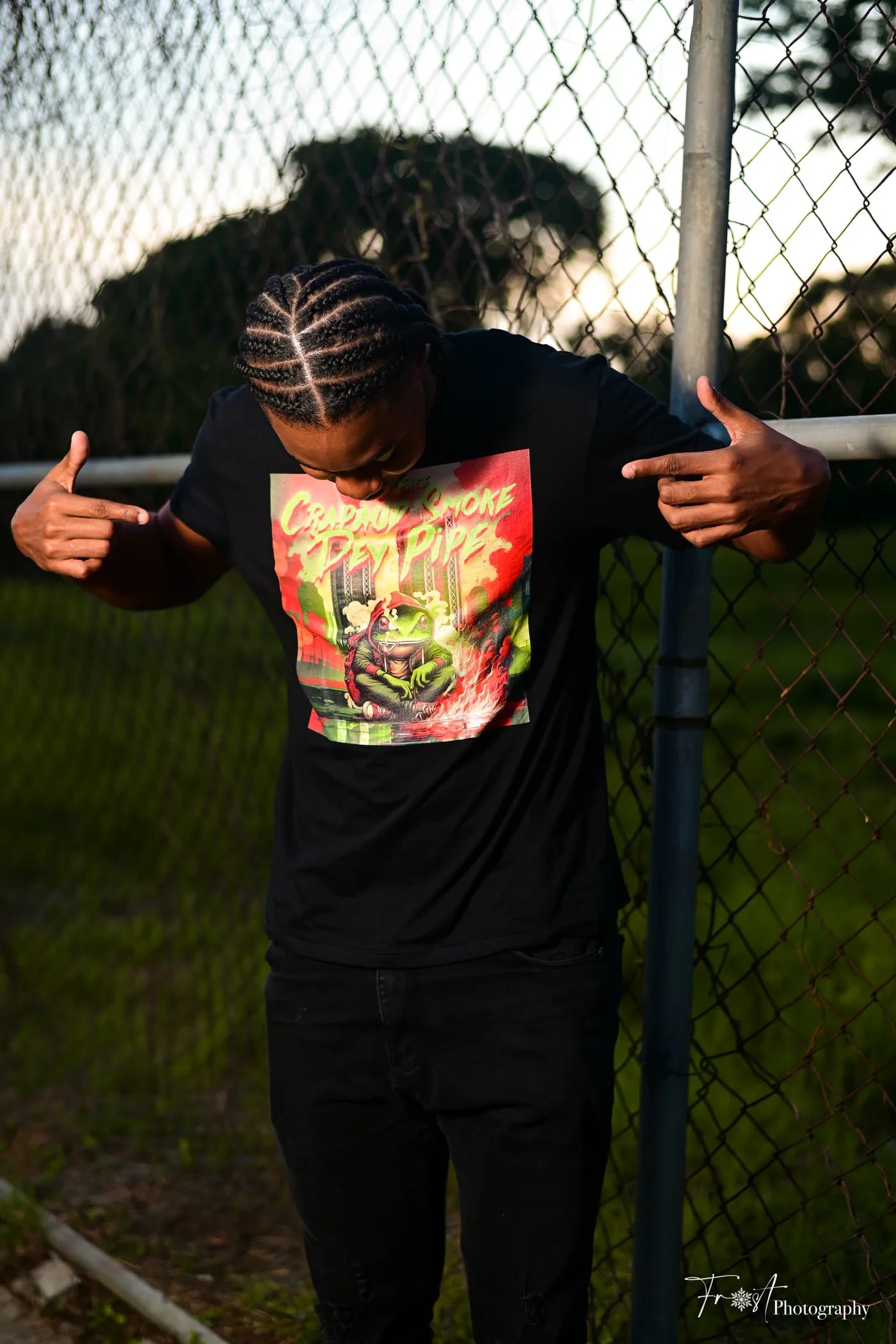 HAMZXL pointing to his Crapaud Smoke Dey Pipe shirt by a chain-link fence.